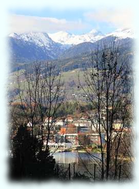 Blick durch die Bäume über den Millstätter See bis Seeboden und schneebedeckte Berge im Hintergrund